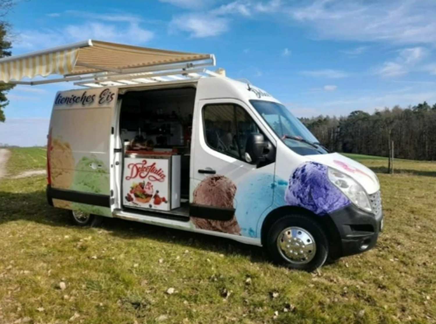 Bayerle - Eiswagen - Ein weißer Eiswagen parkte auf einer grasbewachsenen Wiese unter blauem Himmel. An der Seite prangten bunte Eiscreme-Grafiken, und er hatte eine ausladende, hellbraune Markise.