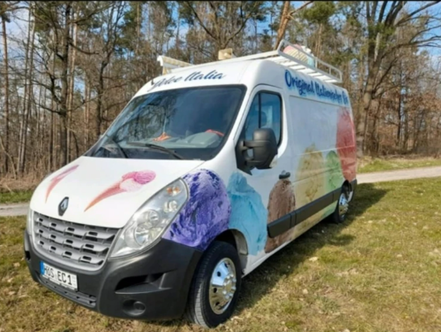 Bayerle - Eiswagen - Ein weißer Eiswagen, geschmückt mit großen, bunten Kugeln Gelato, parkt am grasbewachsenen Straßenrand.