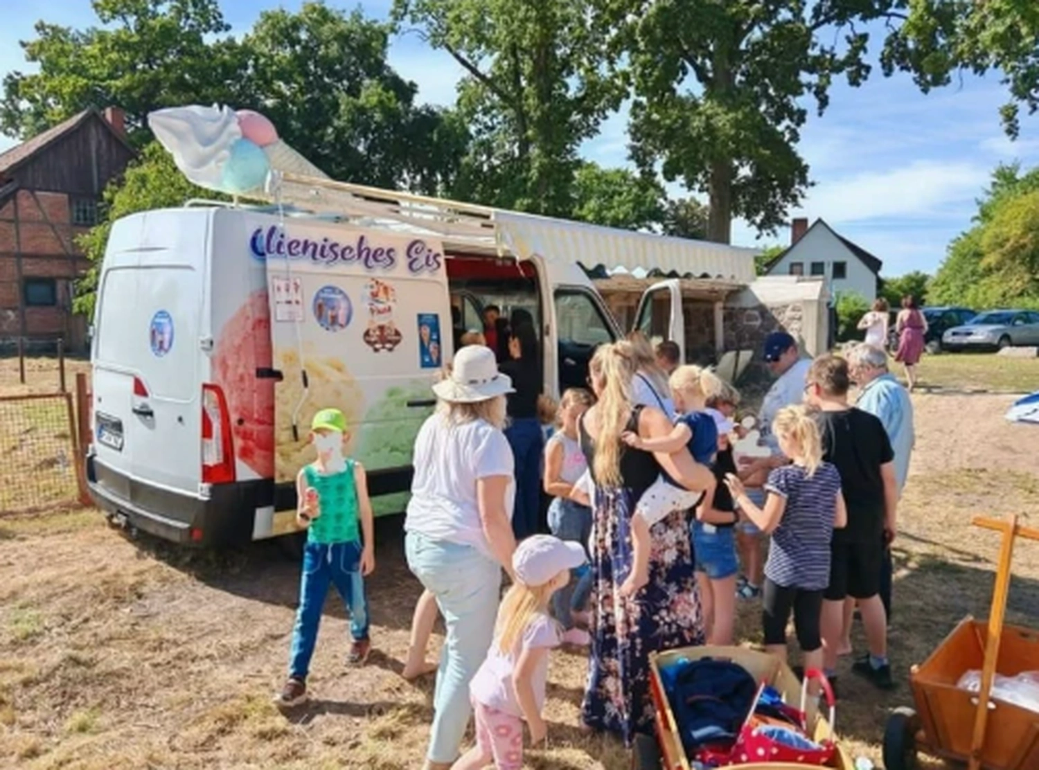 Bayerle - Eiswagen - Ein weißer Eiswagen mit einem großen Eistütenmodell auf dem Dach steht draußen, und an einem sonnigen Tag wartet eine Schlange von Menschen darauf, bedient zu werden.