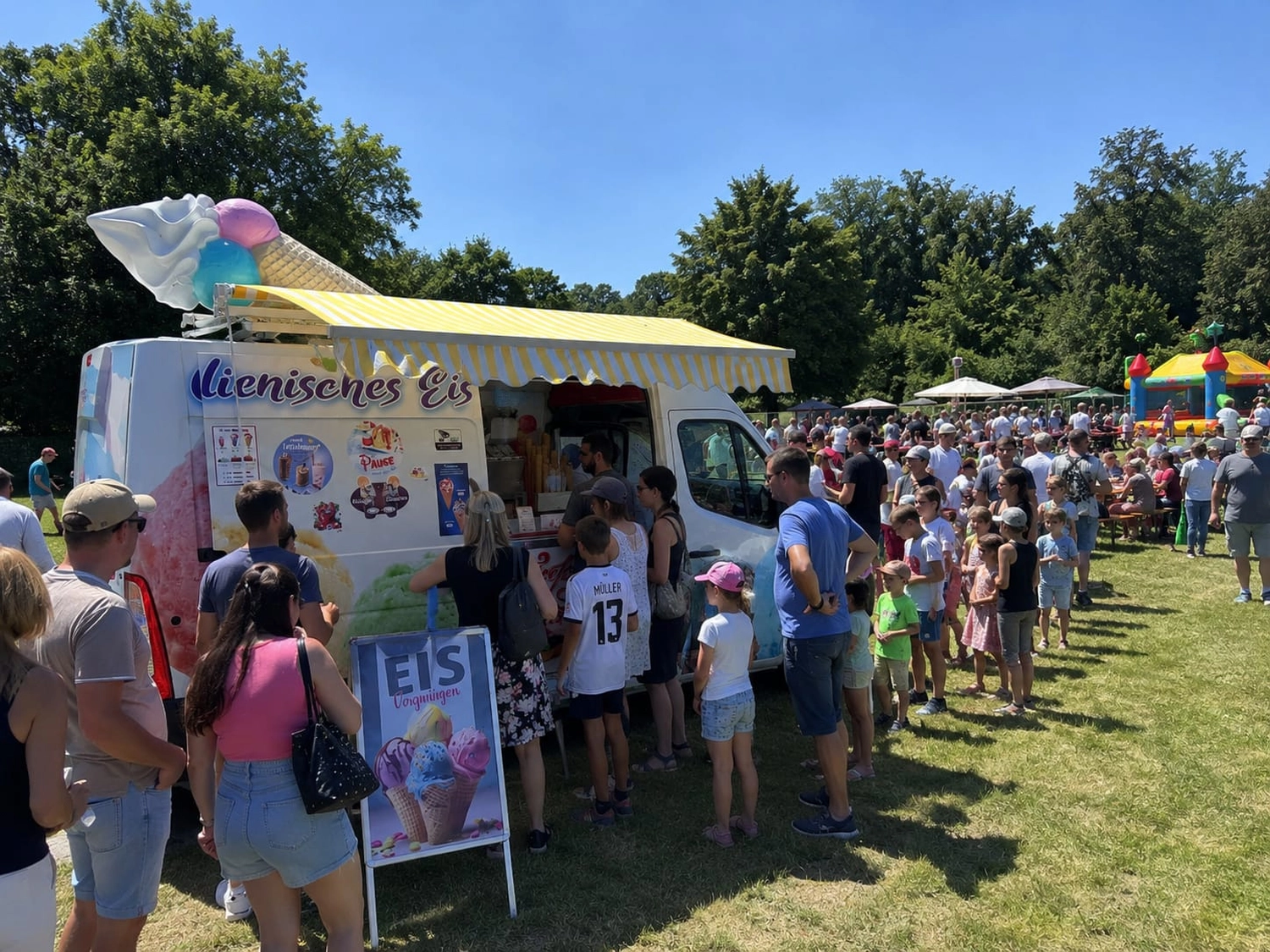 Bayerle - Eiswagen - Eine sonnige Szene im Freien zeigt eine lange Schlange von Menschen, die vor einem weißen Eiswagen der Marke „Lienisches Eis“ auf einer Wiese warten. Der Wagen ist mit einer großen Eistüte auf dem Dach und einer gelb-gestreiften Markise geschmückt, im Hintergrund sind eine bunte Hüpfburg und üppig grüne Bäume zu sehen.